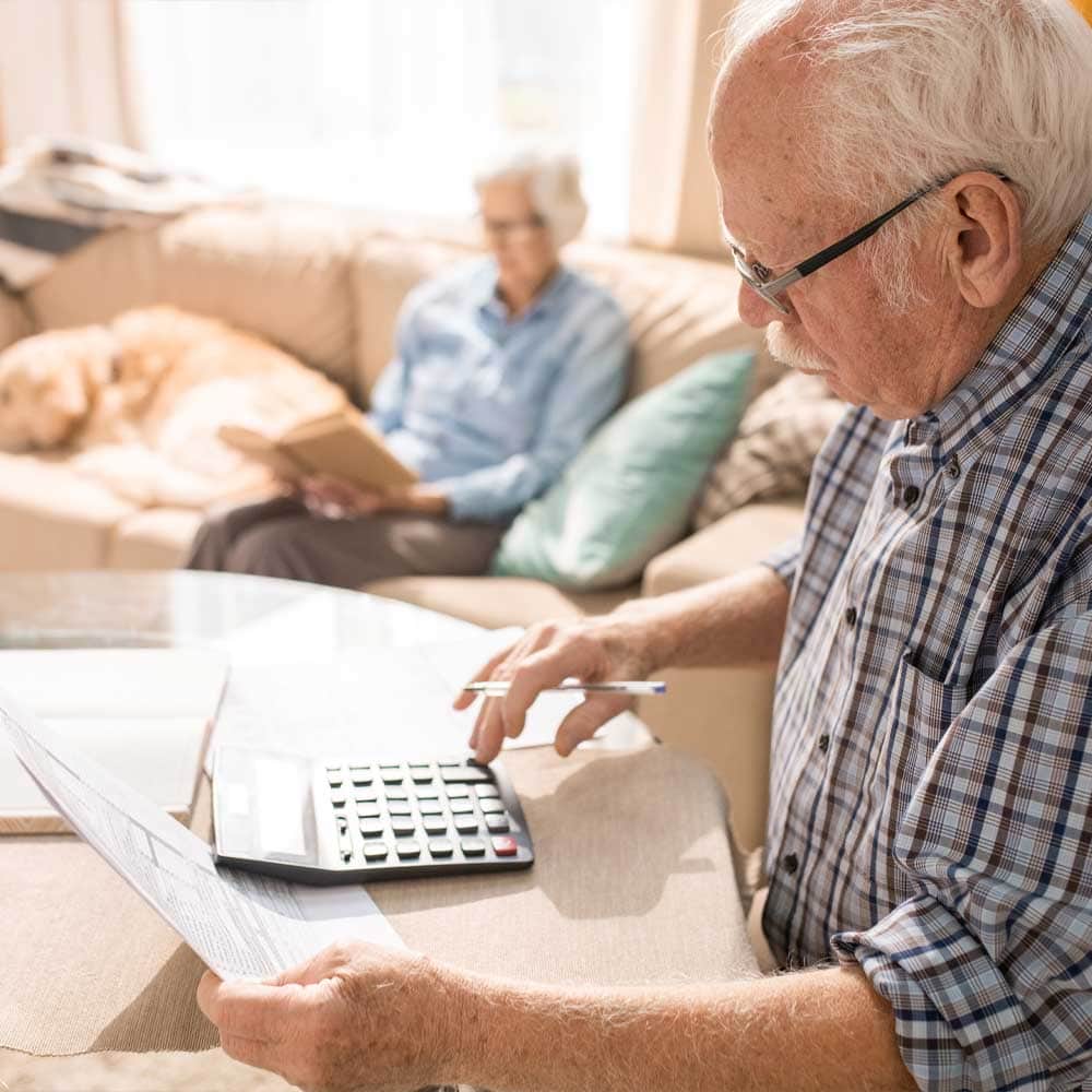 Un senior homme calcule son droit à l'allocation de logement sociale avec sa calculatrice. Sa femme est assise sur le canapé du salon.